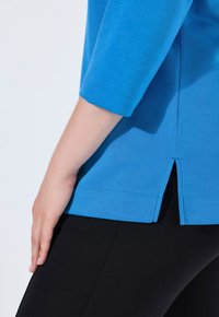 Blusa de algodón azul con mangas de tres cuartos, que presenta una pequeña abertura lateral en el dobladillo, combinada con leggings negros. Textura suave.