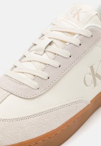 Zapatilla blanca con parte superior de cuero liso y acentos de ante gris claro. Cuenta con puntera redondeada, cordones planos y suela de goma marrón con logotipo.