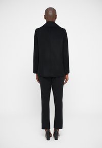 Μαύρο μάλλινο παλτό με ά�νοιγμα κολάρου, δομημένο σχήμα και ευθεία σιλουέτα, συνδυασμένο με μαύρο παντελόνι και ψηλοτάκουνες γόβες.