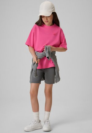 Dziewczyna w różowym t-shircie, szarych krótkich spodenkach, białych trampkach i białej czapce, która związuje szarą bluzę wokół talii.