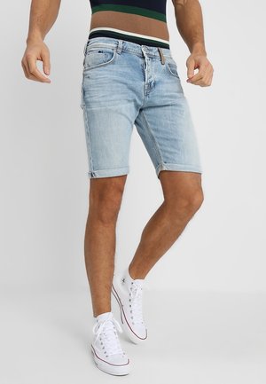 Homme portant un short en jean bleu clair avec ourlets retroussés, debout et chaussant des baskets blanches montantes sur un fond uni.