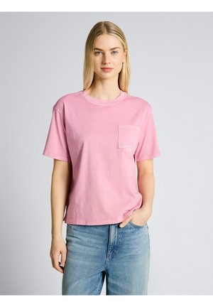 Blondynka w prostym różowym t-shircie z krótkim rękawem i przednią kieszonką oraz jasnoniebieskich dżinsach, stojąca z jedną ręką w kieszeni.