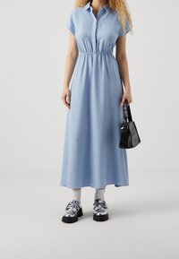 Abito azzurro chiaro a maniche corte, con bottoni e vita elasticizzata, abbinato a una borsa nera e scarpe argento con tacco largo.