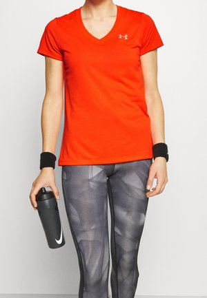 Person iført en lys orange kortærmet top og sorte leggings med mønster, der holder en sort vandflaske med et hvidt logo.
