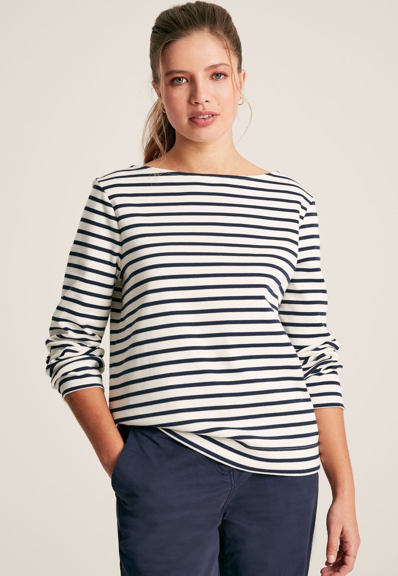 Donna con coda di cavallo che indossa una maglia a maniche lunghe bianca con strisce orizzontali blu navy e pantaloni blu navy, in piedi con una mano in tasca.