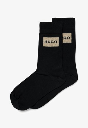 Calze nera in cotone con un bordo a coste e una toppa rettangolare beige con la scritta "HUGO" in lettere nere. Texture liscia.