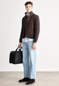 Geacă din piele întoarsă maro și tricotat cu nervuri peste un pulover închis la culoare, cămașă în dungi, blugi evazați deschiși la culoare și ghete negre până la gleznă, ținând o geantă neagră de voiaj.