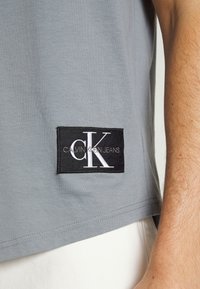 Camisa gris con un parche negro de Calvin Klein Jeans en el lado inferior, junto a un brazo desnudo de una persona y pantalones blancos.