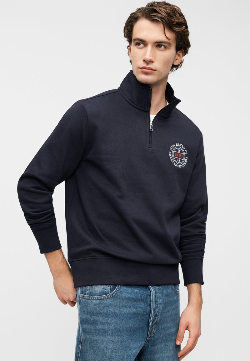 Felpa blu navy scuro con mezza cerniera, realizzata in un tessuto morbido, con polsini e orlo a costine, e un dettaglio logo circolare sul petto a sinistra.