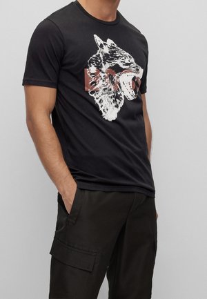 Mann trägt schwarzes T-Shirt mit Grafik eines brüllenden Wolfes und dunkle Cargoshorts, steht mit einer Hand in der Tasche vor einfachem Hintergrund.