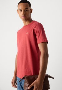 Un joven que lleva una camiseta roja sostiene una bolsa de aseo de cuero marrón frente a un fondo claro y liso.