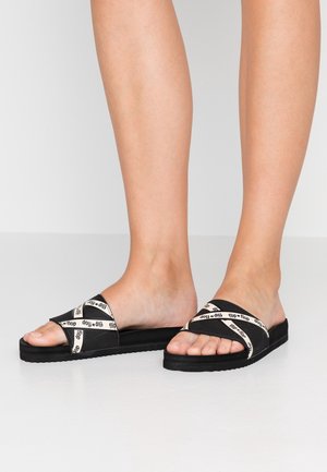 Czarne klapki na płaskiej podeszwie z paskami skrzyżowanymi, ozdobione kremowym nadrukiem z logo. Płaska podeszwa z teksturowanej gumy zapewniająca przyczepność i komfort.