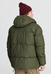 Zielona kurtka puchowa z kapturem, z pikowanymi sekcjami, gładką fakturą i regulowanymi bocznymi trokami. Noszona z bordową czapką.