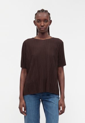 Femme aux cheveux tressés portant un haut plissé marron foncé à manches courtes et un jean bleu, debout devant un fond clair et uni.