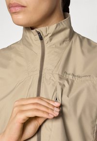 Light beige, zippered performance jacket with high collar, lightweight fabric, and a discreet chest pocket. Brand logo visible on the chest.