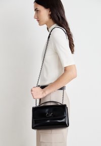 Giovane donna di profilo che tiene una piccola borsa nera con tracolla a catena, indossando un top bianco aderente e pantaloni beige, su uno sfondo chiaro.