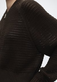 Gros plan d'une personne portant un pull en maille texturée marron foncé avec une fermeture éclair à l'encolure et des manches longues.