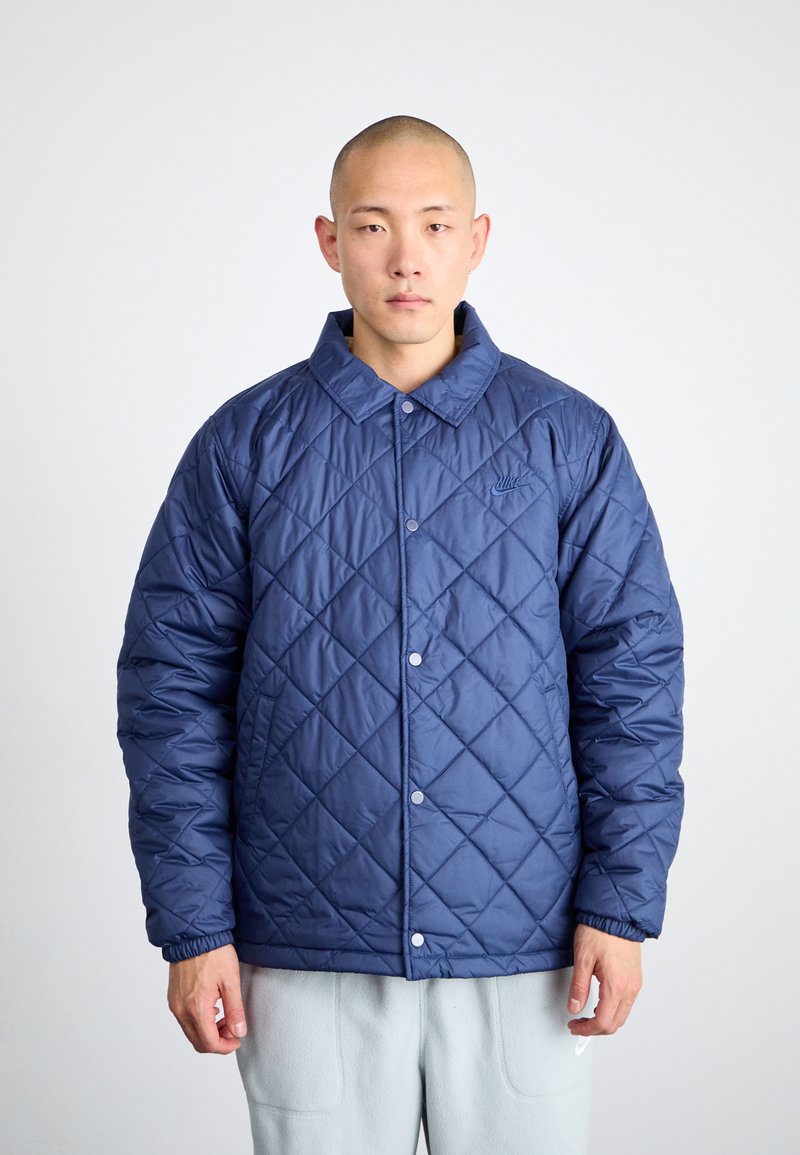 Veste matelassée bleu marine avec fermeture à boutons, col montant et poignets élastiques. Présente un petit logo sur la poitrine gauche.