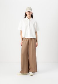 Camisa blanca de manga corta con un cierre de botones, combinada con pantalones amplios de color marrón claro. El conjunto se complementa con una gorra beige y zapatillas blancas.