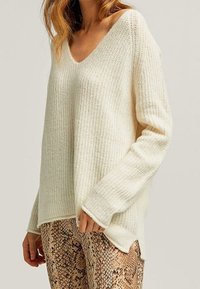 Femme portant un pull en maille côtelée couleur crème à col en V et un pantalon à motif serpent marron, posant avec une main près de la hanche.