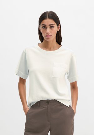 Frau mit dunklen Haaren, die ein weißes kurzärmeliges Hemd mit Brusttasche und braune Hosen trägt, steht mit den Händen in den Taschen vor einem schlichten Hintergrund.