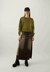 Jovem mulher vestindo um sweatshirt verde-oliva, boné preto, brincos de argola, saia longa castanha escura com uma abertura lateral e botas de plataforma pretas.