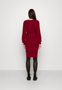 Femme portant une robe en tricot bordeaux cintrée avec des manches longues, des collants noirs et des chaussures noires, se tenant face à un mur blanc uni.