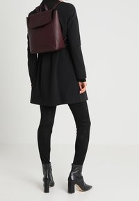 Zaino in pelle bordeaux con chiusura a patta, dotato di spallacci regolabili, una texture liscia e un design strutturato, indossato con abiti neri.