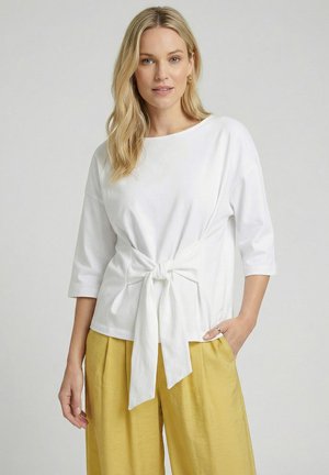 Blonde Frau trägt ein weißes Dreiviertelärmeliges Oberteil mit vorderer Bindung und lockere senfgelbe Hose, steht vor einem einfachen Hintergrund.