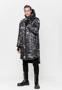 Czarna kurtka puchowa z białym wzorem plam, z wysokim kołnierzem i zapięciem na zamek. Nosi ją osoba w czarnych spodniach i butach. Gładka, błyszcząca faktura.