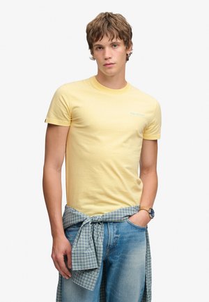 Jonge man die een lichtgeel T-shirt draagt met een geruit overhemd om zijn middel geknoopt en een blauwe spijkerbroek, staand met één hand in zijn zak.