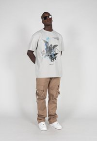 Grå grafisk t-shirt med blå og sorte designs, parret med beige cargo bukser. Modellen bærer sorte solbriller og hvide sneakers.