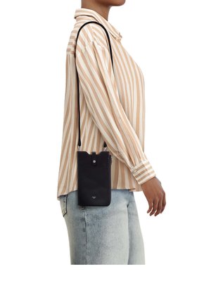 Frau trägt beige-weiß gestreiftes Hemd und hellblaue Jeans und trägt eine kleine schwarze Umhängetasche mit langem Riemen.