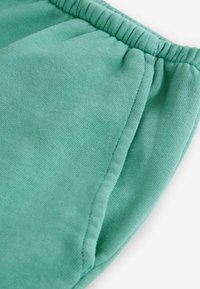 Shorts sportivi color teal realizzati in tessuto morbido, con vita arricciata e tasche laterali, presentano una texture liscia e un design casual.