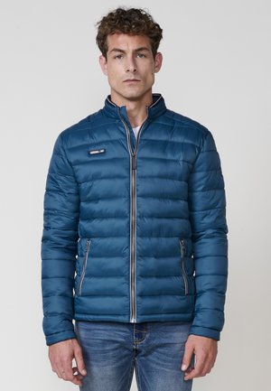 Chaqueta acolchada azul con cuello alzado, textura de acolchado vertical, bolsillos con cremallera y un acento de logo metálico en el pecho.