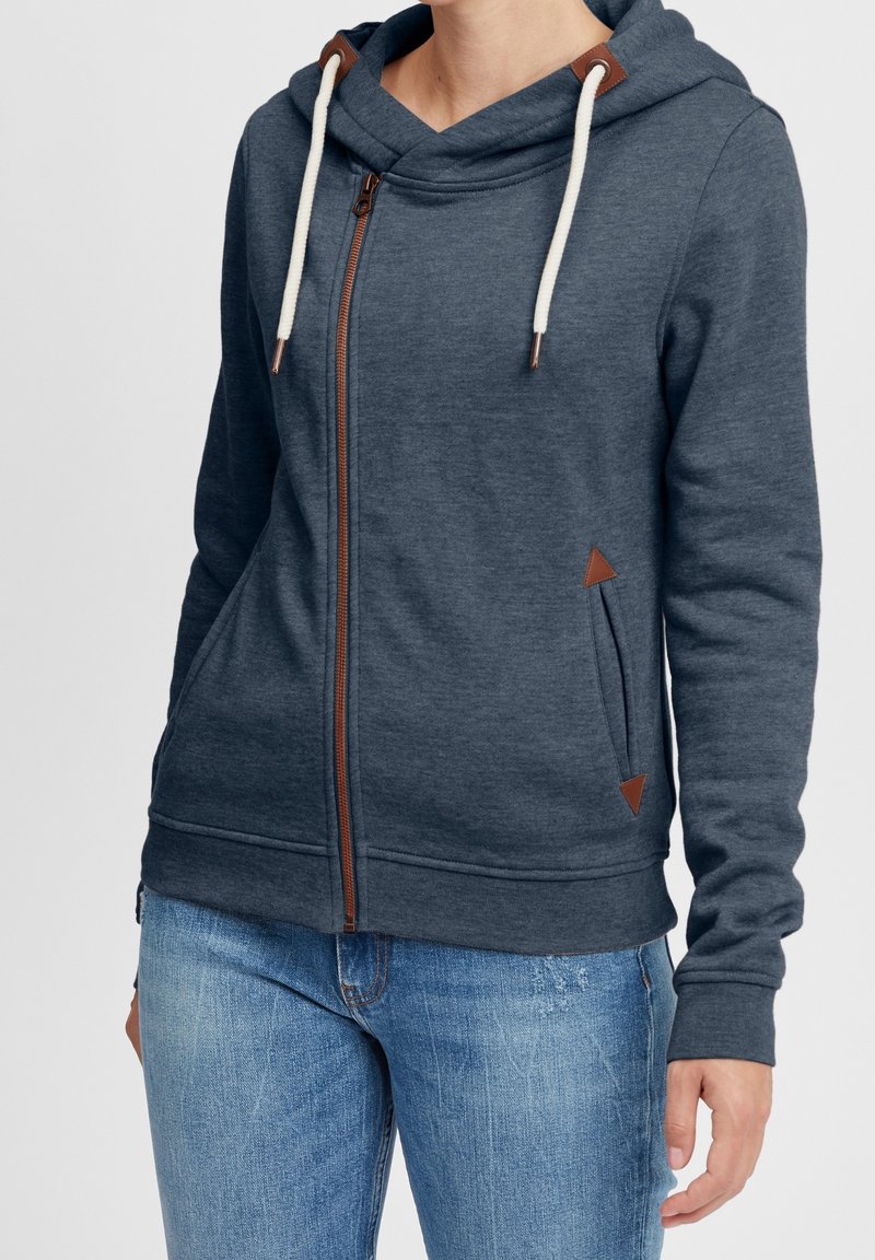 Sudadera azul con cremallera frontal, cordón ajustable en la capucha y acentos de cuero marrón. Cuenta con bolsillos laterales con detalles triangulares.