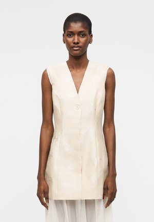 Sötét bőrű, hátrafogott hajú nő ujjatlan, krémszínű, gombos mellényt visel, enyhe virágmintával, fehér háttér előtt áll.