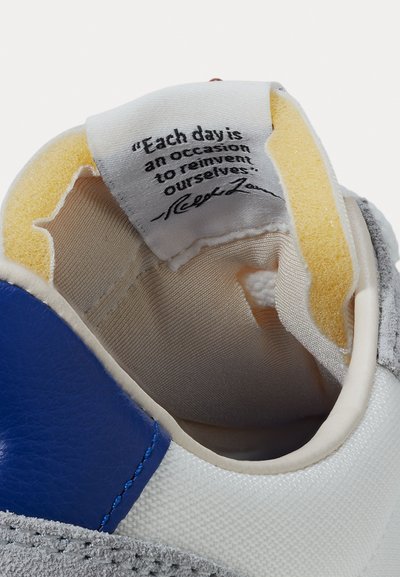 Λευκό εσωτερικό αθλητικού παπουτσιού με μπλε και γκρι λεπτομέρειες, υφή σκουρόχρωμου σουέτ, και κίτρινη αφρώδη επένδυση. Περιλαμβάνει ετικέτα με εκτυπωμένη εμπνευσμένη φράση.