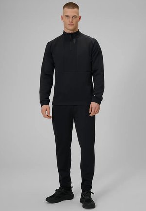 Mężczyzna stojący, ubrany w czarną bluzę na suwak i dopasowane czarne spodnie oraz czarne buty sportowe, na jednolitym szarym tle.