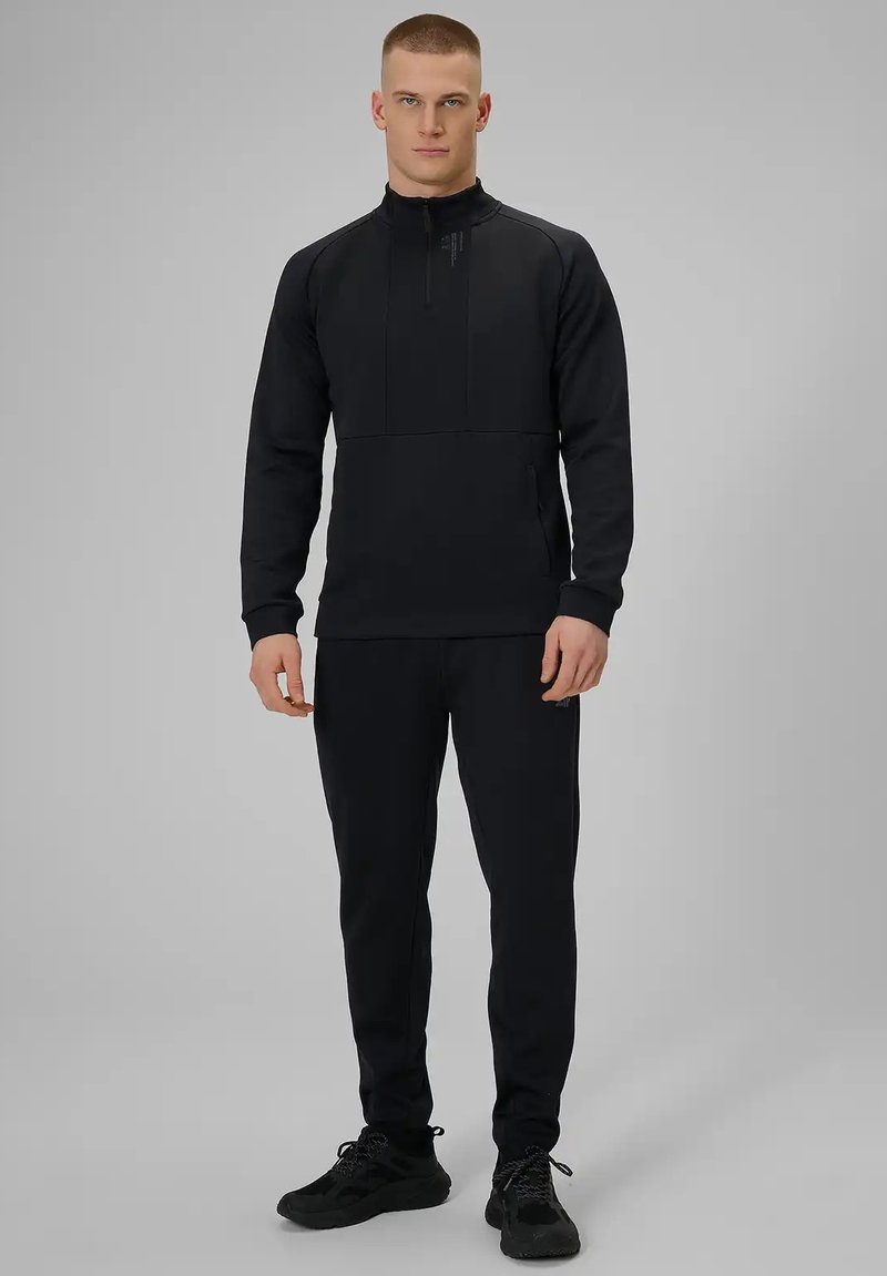 Homme debout portant un sweat à capuche noir zippé et un pantalon noir assorti avec des baskets noires, sur un fond gris uni.