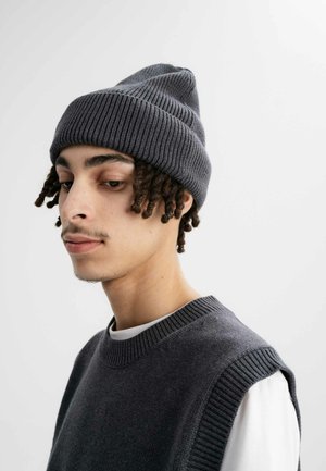 Grijze geribbelde gebreide beanie met een puntige top, gedragen over een grijze mouwloos sweater. De textuur is glad met verticale breipatronen.