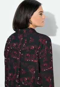 Camicia nera con un motivo astratto rosso, dotata di colletto e schiena con bottoni, realizzata in un tessuto liscio con una leggera lucentezza.