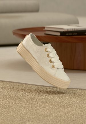 Baskets blanches brodées de fleurs avec une semelle épaisse beige et de grands lacets en satin, flottant à l'intérieur près d'une table basse en bois avec des livres.