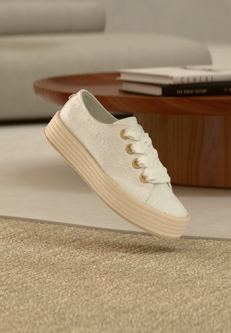 Λευκό αθλητικό παπούτσι κεντημένο με λουλούδια, με χοντρή μπεζ σόλα και μεγάλα σατινέ κορδόνια, αιωρείται σε εσωτερικό χώρο κοντά σε ξύλινο τραπεζάκι καφέ με βιβλία.