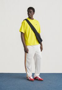 Νεαρός άνδρας που φοράει φωτεινό κίτρινο T-shirt Adidas, λευκό παντελόνι με κόκκινες, κίτρινες και μαύρες ρίγες, κόκκινα αθλητικά παπούτσια Adidas και μαύρη τσάντα χιαστί.