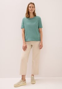 Mujer de pie contra un fondo pálido, vistiendo una camiseta de manga corta teal con texto, pantalones beige de pierna ancha y zapatillas beige.