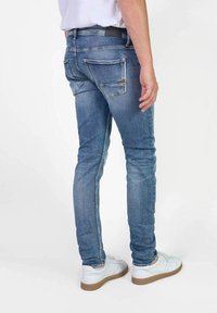 Jeans in denim blu con taglio slim, dotati di due tasche posteriori e vita media, indossati con sneakers bianche.