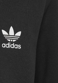 Černá bavlněná látka s hladkou texturou. Na ní je velké bílé logo Adidas s třemi pruhy nad názvem značky.