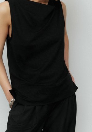 Mouwloze zwarte top met gedrapeerde halslijn en bijpassende zwarte broek, persoon die een zilveren kettingarmband draagt met handen in de zakken.