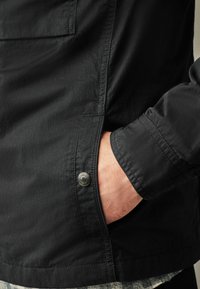 Veste noire avec une finition lisse, dotée de poches latérales, d'accents en bouton et d'une coupe décontractée. Manches texturées et détails à l'ourlet visibles.